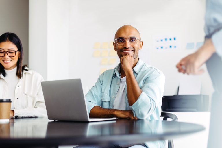 Male designer smiling at the camera while sitting at a table with his colleagues. Black business man having a meeting with his team in an office.